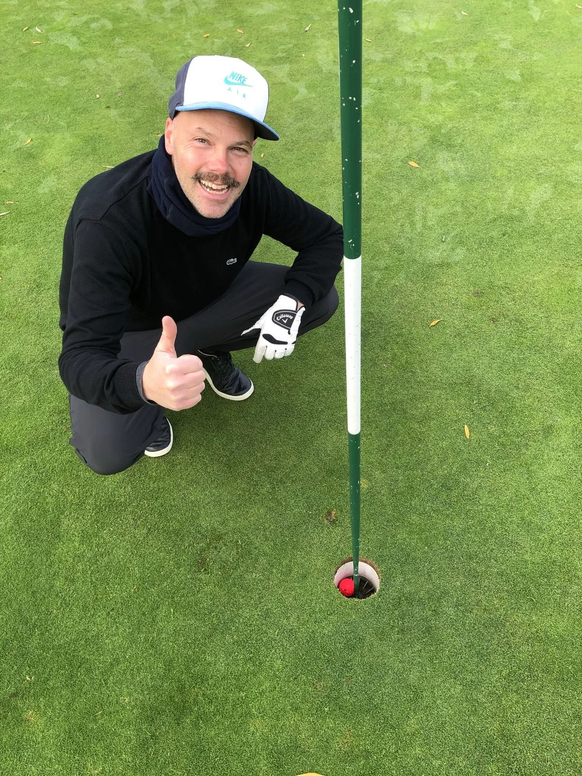 Björn Rosenthal kneeling beside the hole giving a thumbs up, the red ball sitting in the cup